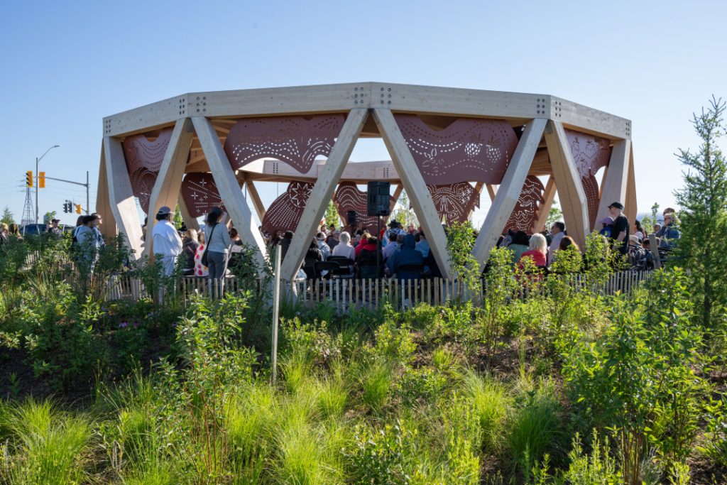 People gathered in and around drum structure in Biidaasige Park, surrounded by fencing and grass.