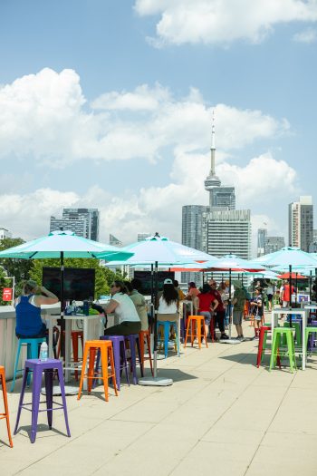 People sit at high-top tables with colourful stools and umbrellas. There are TV screens showing a soccer game. City skyline in the background.