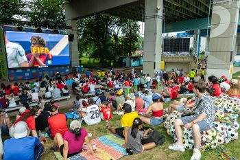Crowd sits on a grassy hill underneath Gardiner Expressway watching a large TV screen showing FIFA soccer player.
