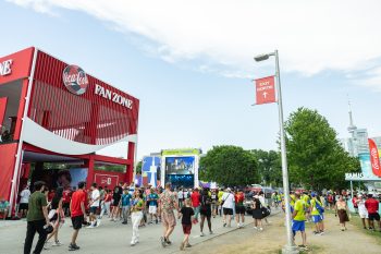 Crowds walk through a festival grounds. Building on left says Coca-Cola Fan Zone