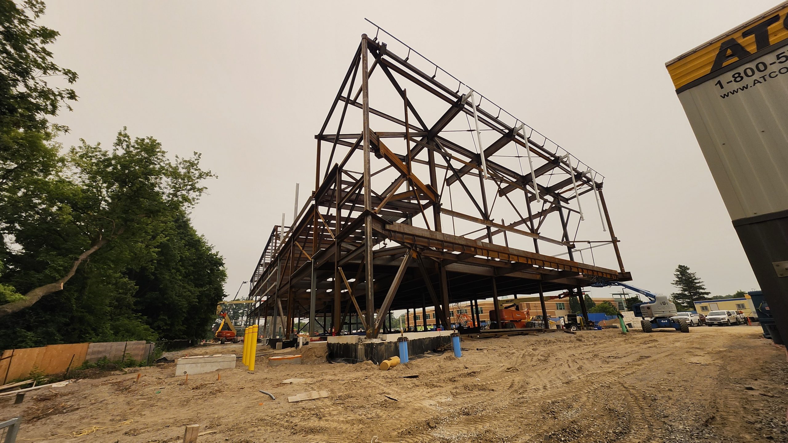 View of the steel structure for the new community recreation centre in progress. The multi-storey structure is supported by large concrete blocks embedded in the dirt ground. Machines such as lifts and excavators are visible around the construction site.