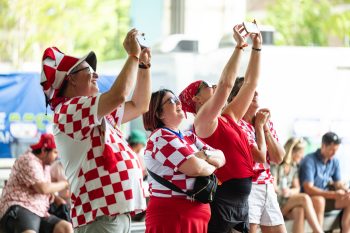 Four people stand in the foreground, smiling and holding mobile phones up to the right. Three are wearing Croatia soccer jerseys. In the background, people are seated.