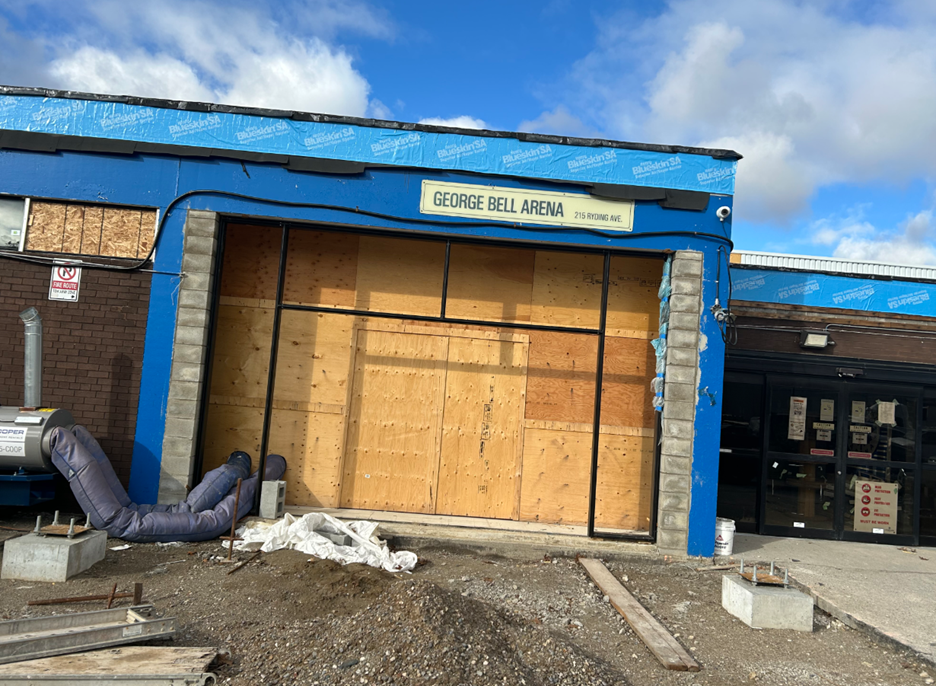 Exterior of George Bell Arena at 215 Ryding Avenue, showing a boarded-up entrance under construction with plywood panels, blue weather barrier wrap, and construction materials on the ground.
