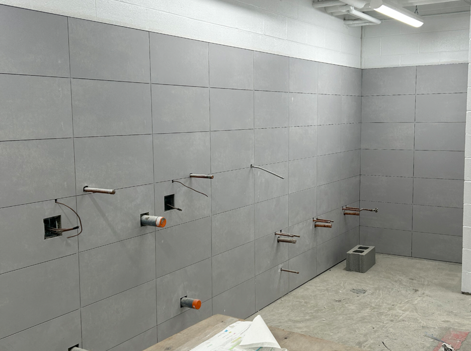 Partially completed grey tiled wall in an interior space with several plumbing pipes and electrical conduits protruding from the surface, alongside a concrete block on the floor.