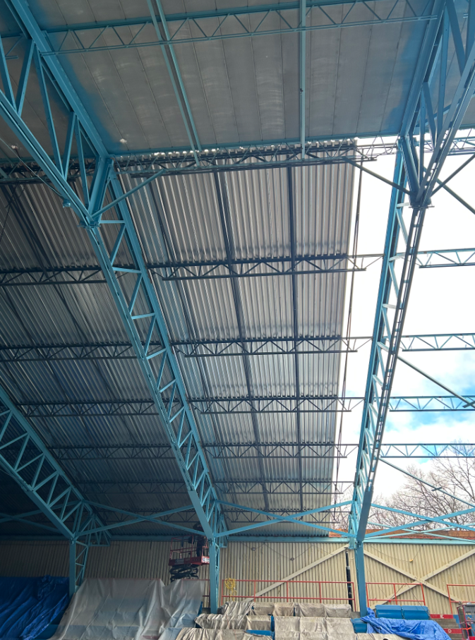 Interior view of a large open-air structure's roof under construction, showing exposed blue steel trusses and metal roof decking against a partly cloudy sky.