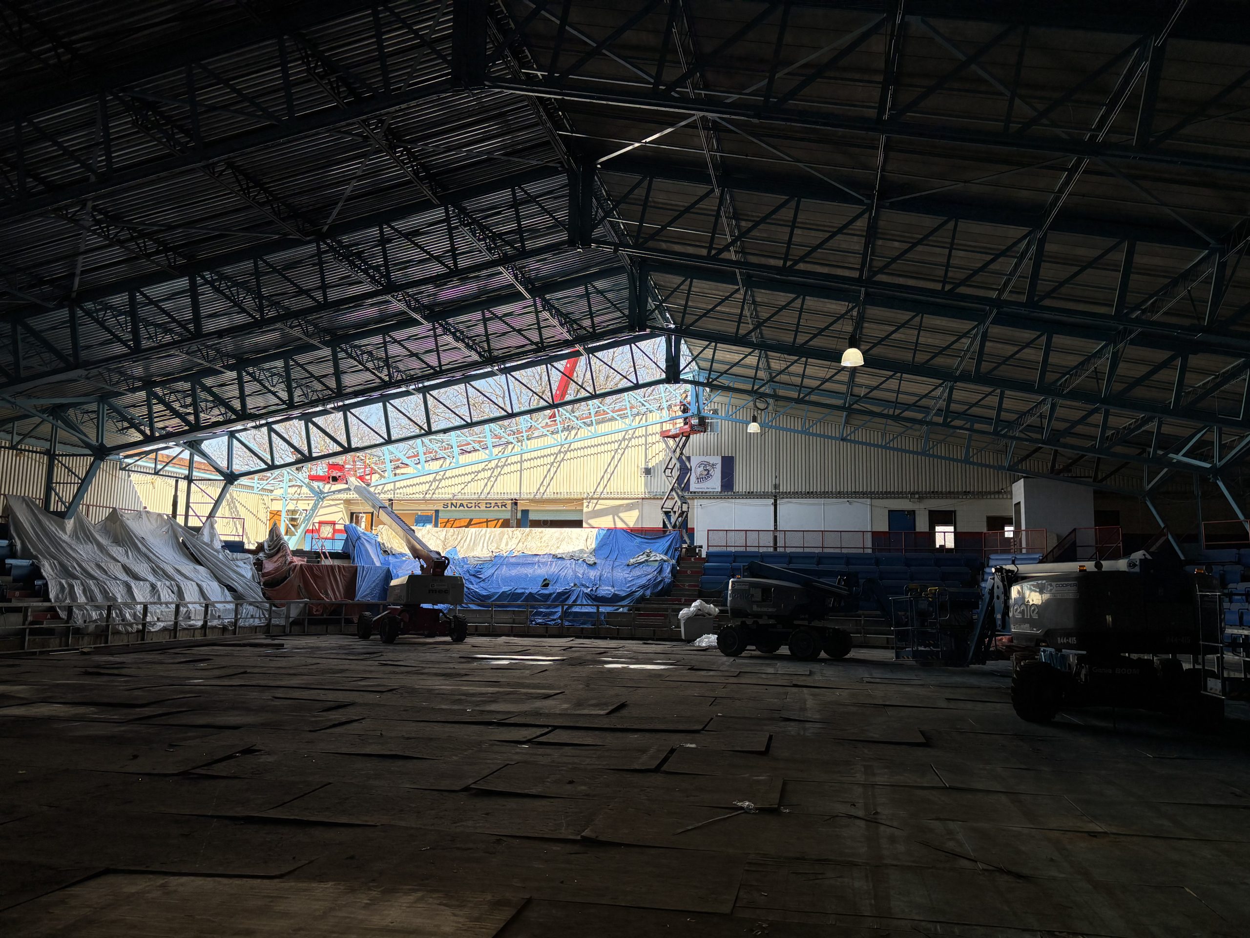 The rink in George Bell Arena under construction. Steel beams span the ceiling and a section of the roof is open, letting in natural light. The rest of the space is dim. Construction equipment and materials are piled on one end.