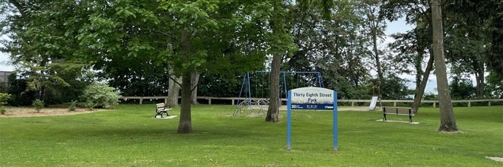 image of the current playground at Thirty-Eight Street Park, featuring a grassy park space with a climbing dome, a swing set and a slide sitting on a sandy surface.