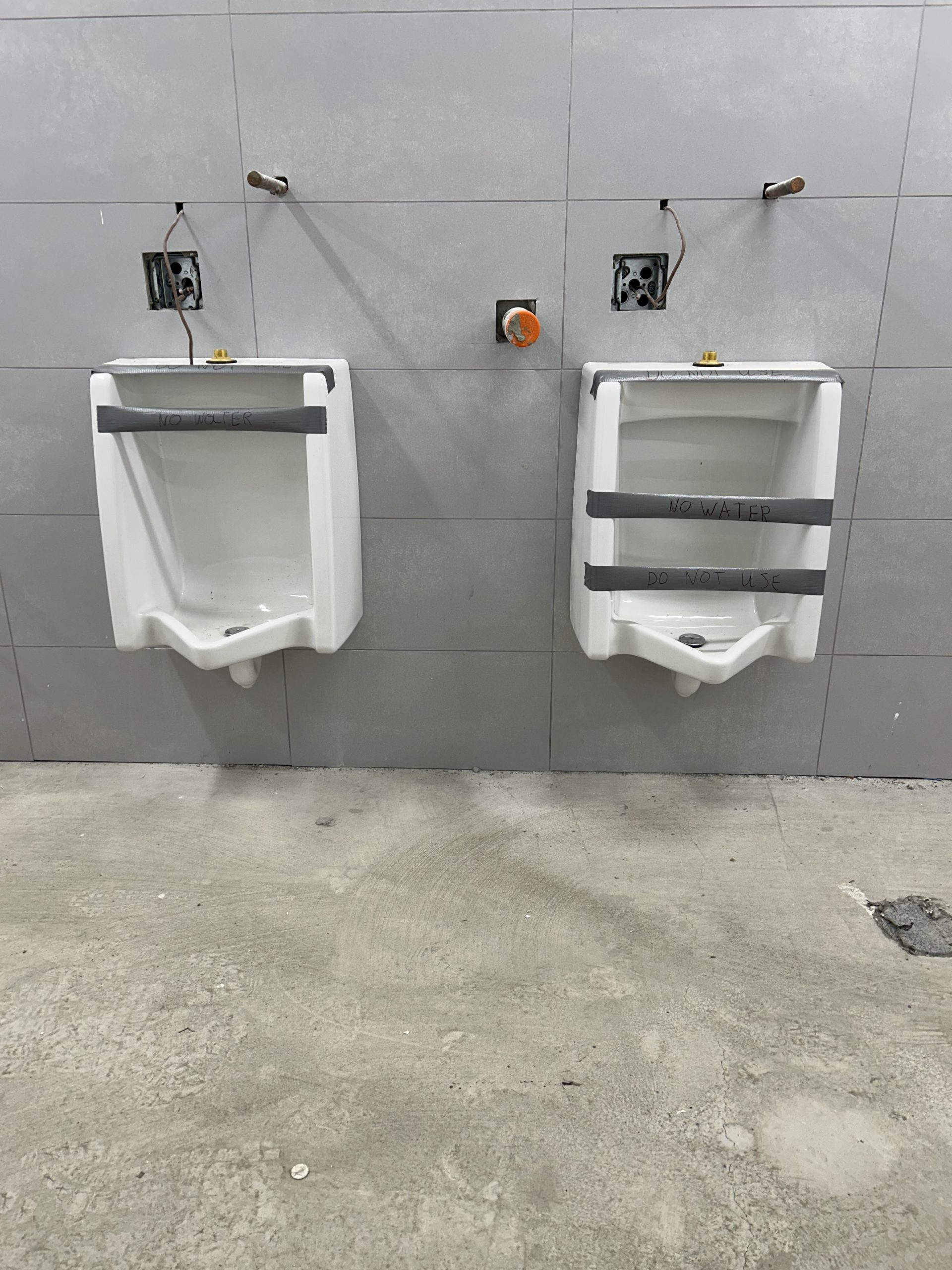 Two taped off urinals installed against a grey tiled wall. Plumbing and electrical connections remain unfinished above the urinals. The concrete floor is unfinished.