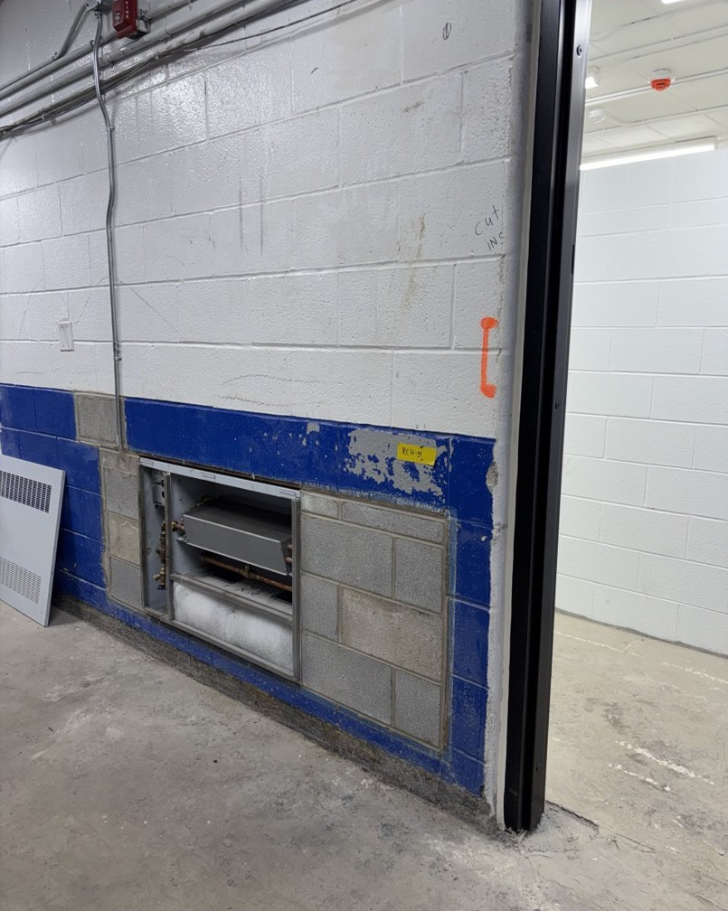 View of a block wall in George Bell Arena. The lower half of the wall is partially painted in blue and has an opening where radiator parts have been installed. Electrical wiring runs along the top of the wall.