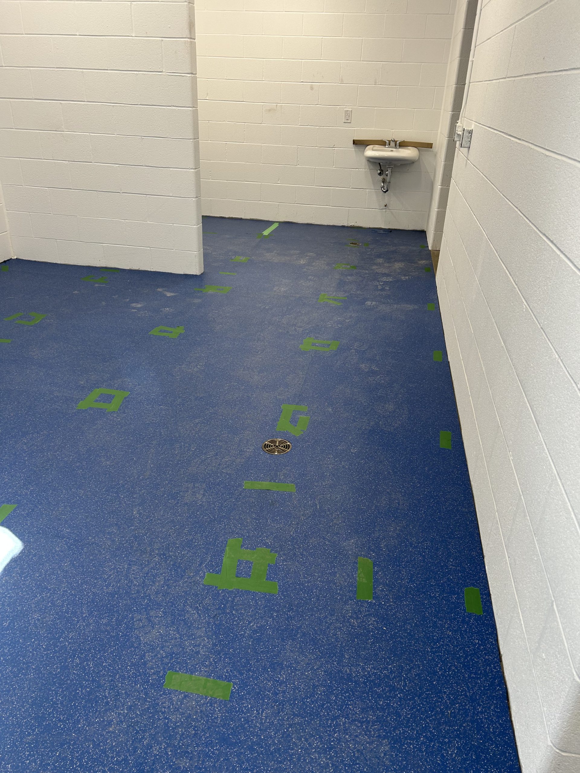 A dressing room in George Bell Arena with white block walls and blue speckled floors. Strips of green tape mark spots on the blue speckled floor. A small sink is mounted on the wall at the far end.