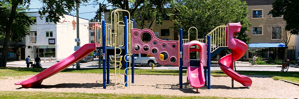 Image of the existing playground at Allan A. Lamport Stadium Park, featuring a main play structure with red slides, yellow climbing ladders, bridges and interactive play panels set on a sandy surface. There are trees surrounding the playground, with grassy open space and nearby buildings visible in the background.