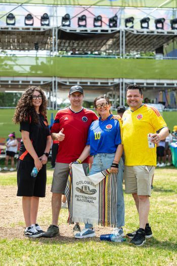 Four people wearing soccer jerseys pose for the camera. One holds Colombia Patrimonio Nacional banner. In the background is festival tiered seating and festival-goers.