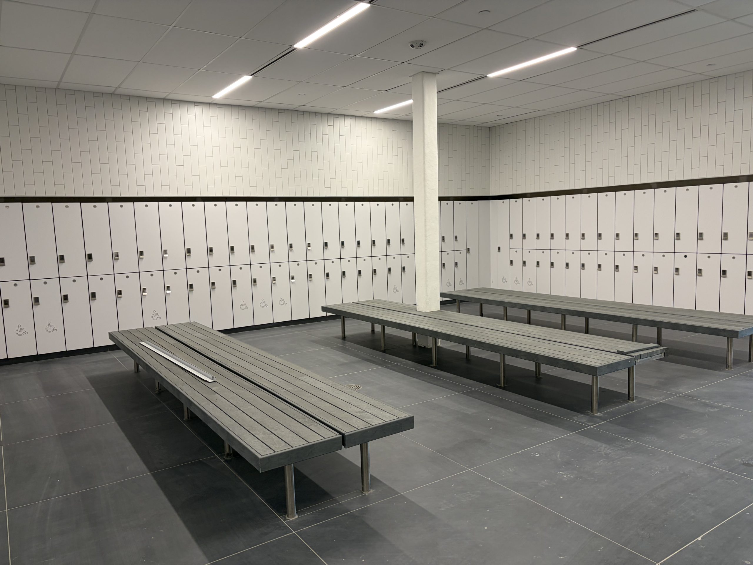 View of the change rooms, showing three long benches surrounded by white lockers along the walls.