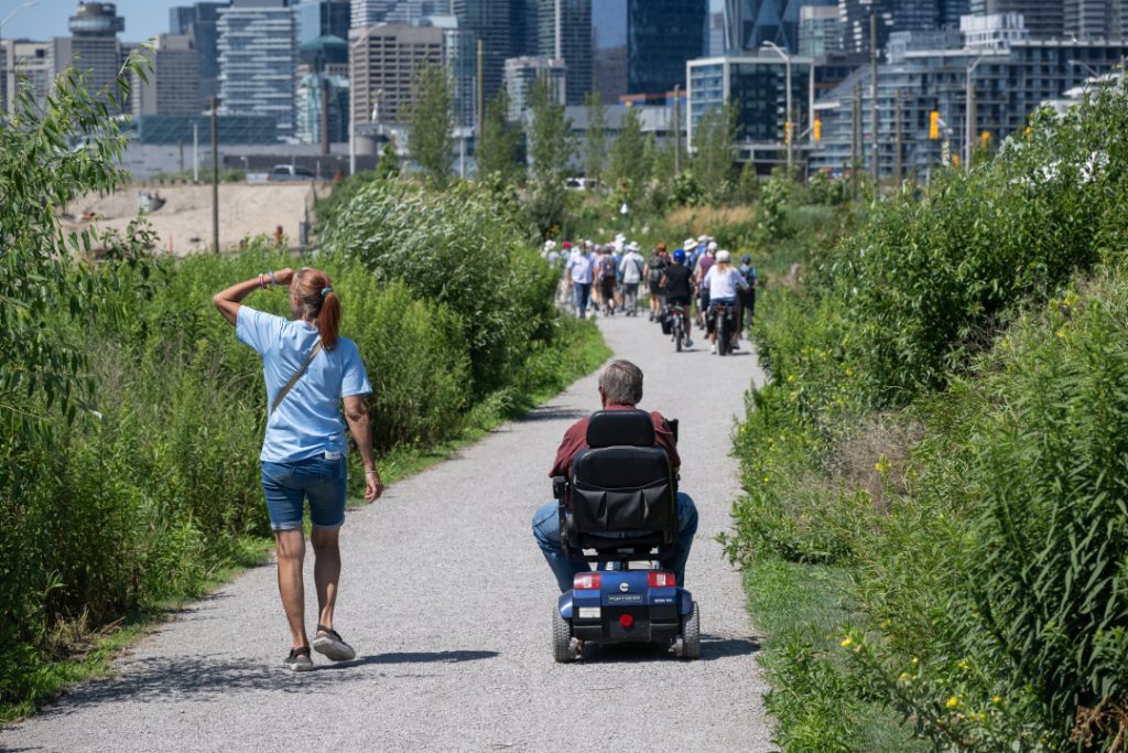 A woman walking alongside a man riding motorized scooter on a trail in Biidaasige Park with grass on both sides and city buildings in background.