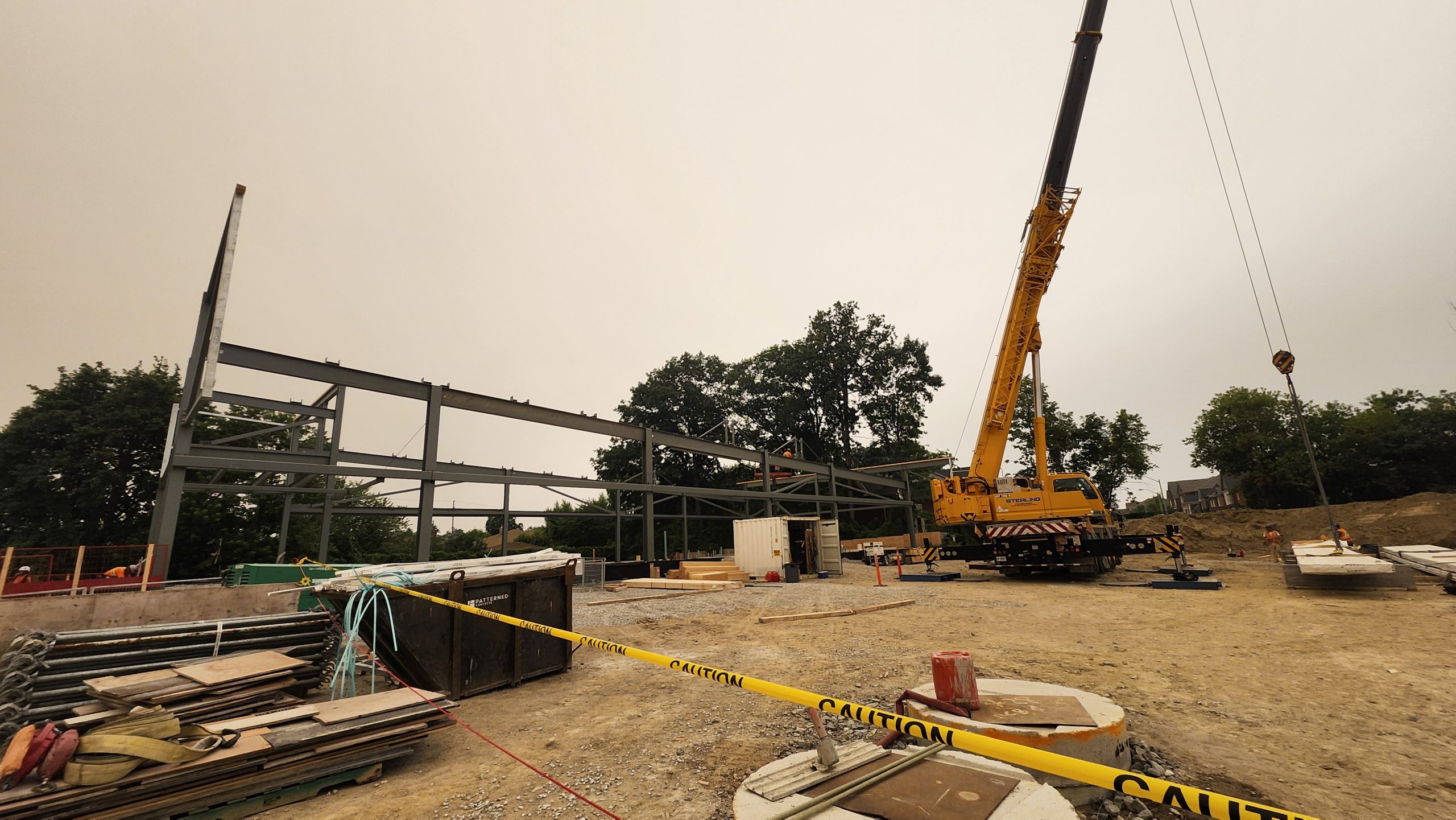 View of the construction site for the new community recreation centre, showing a building’s structural framework in progress. A tall crane at the centre of the site is hooked onto a large slab. Piles of construction materials are scattered around, while trees line the background.
