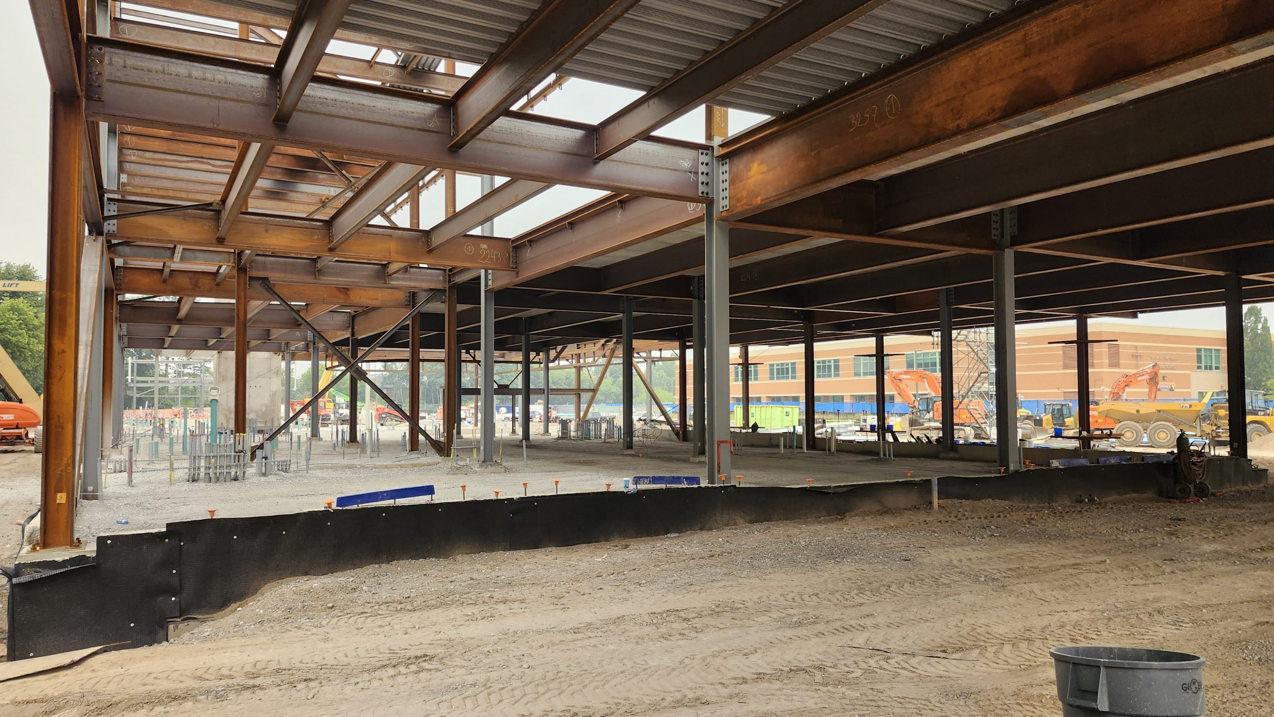 View of the construction site from under the steel structure for the new community recreation centre, made up of beams, supporting columns and metal panels. Through the open sides of the structure, construction equipment is visible parked on the gravel ground.
