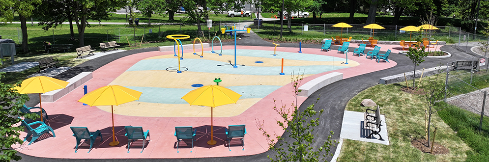 Aerial view of a splash pad in a park. Water features are situated in the centre on a colourful concrete surface, surrounded by lounge chairs and patio-style tables with umbrellas. The colour scheme of the splash pad is yellow, orange, blue and teal.