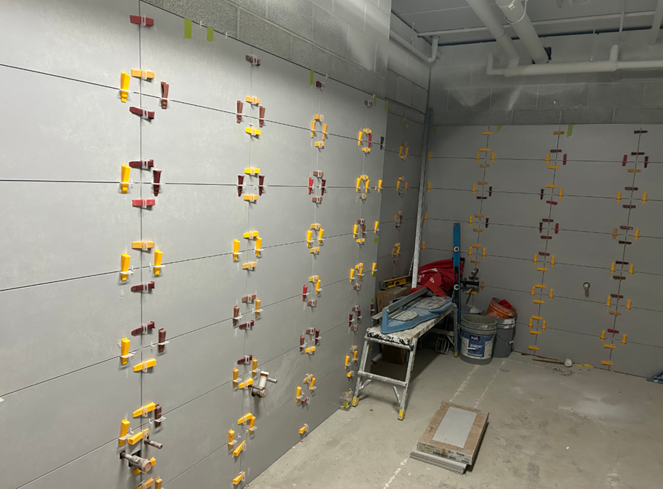 Interior construction site with grey tile spacers and leveling clips installed across three unfinished tiled walls, a ladder, tile-cutting tool, and building materials on the floor.