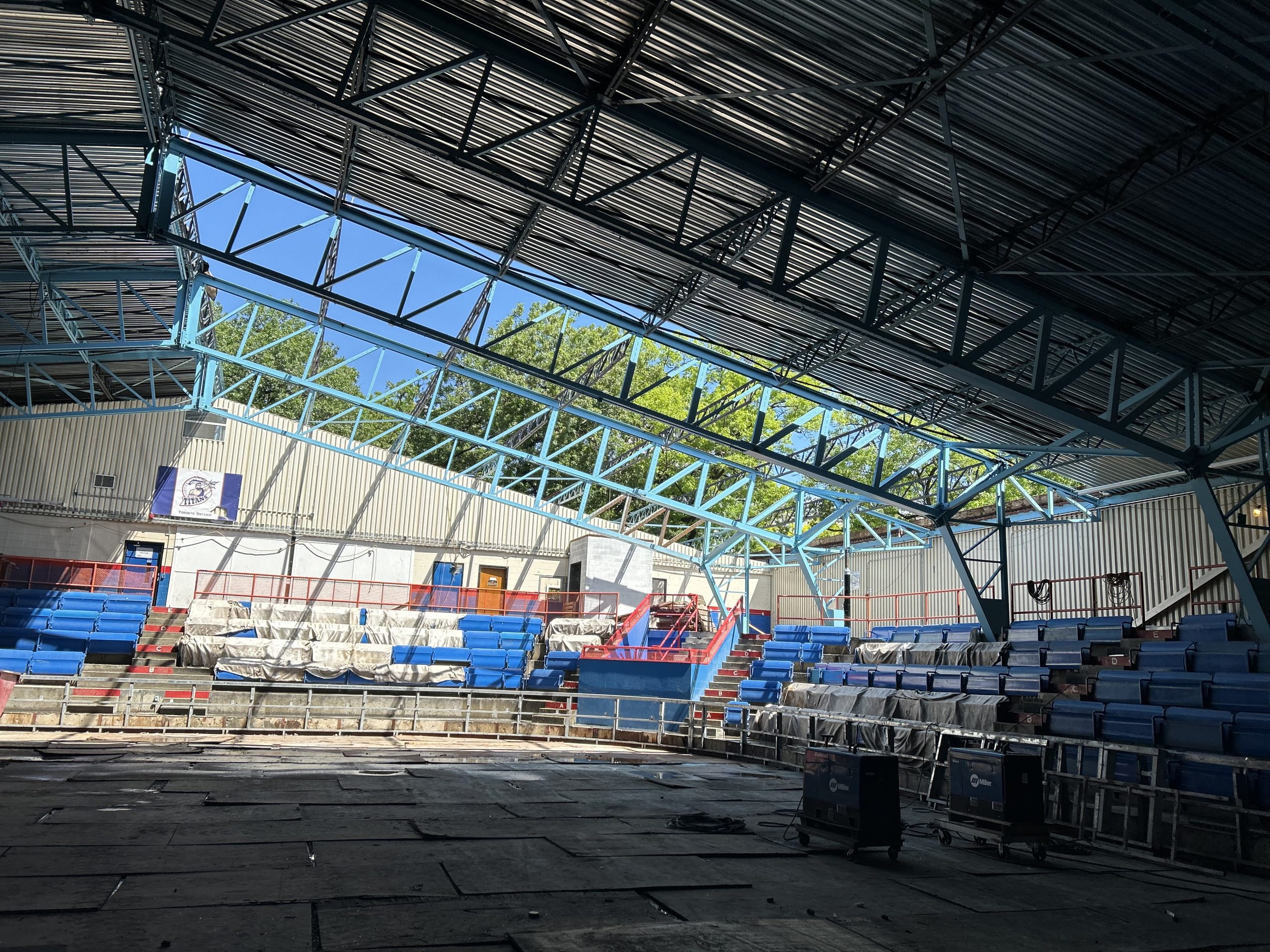 The rink in George Bell Arena under construction, showing the ongoing roof installation. Blue steel trusses support the roof, which is mostly in place except for one open section. Blue bleachers surround the unfinished rink surface where construction equipment is visible.