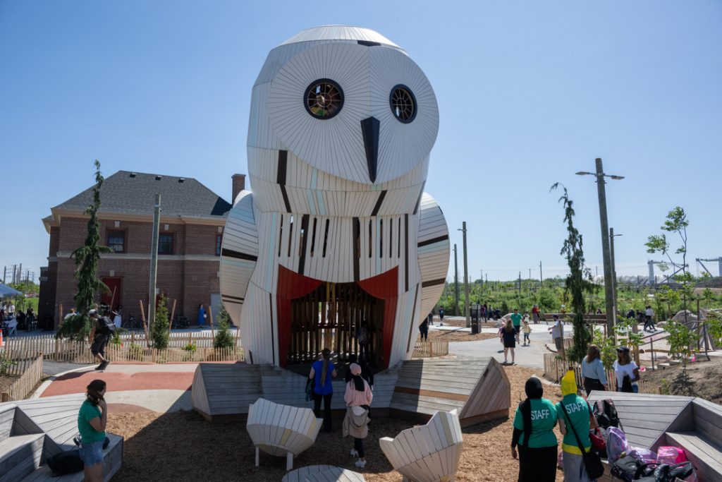 Visitors at Biidaasige Park looking up at snowy owl structure with fire hall in background.
