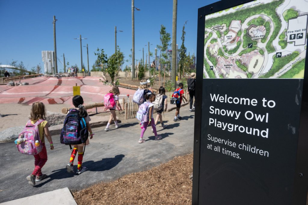 Children walking on trail in Biidaasige Park, near black sign reading Welcome to Snowy Owl Playground, Supervise children at all times.