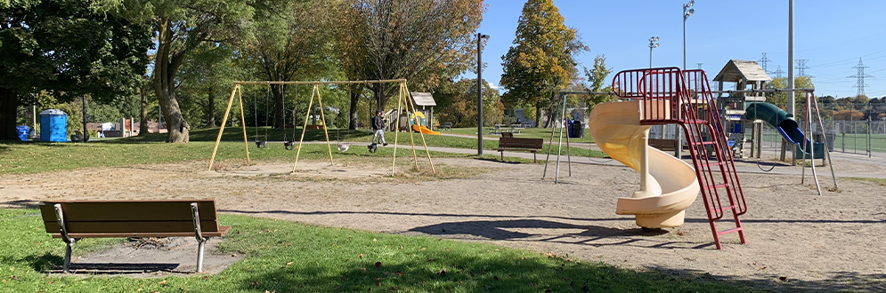 Photo image of the current playground at McNicol Park showing play features on a sandy ground surface. From left to right, this playground includes two benches, a swing set with four toddler swings, a junior play structure with a spiral slide in front, a senior play structure with a curved slide at the back and another swing set with four belt swings.