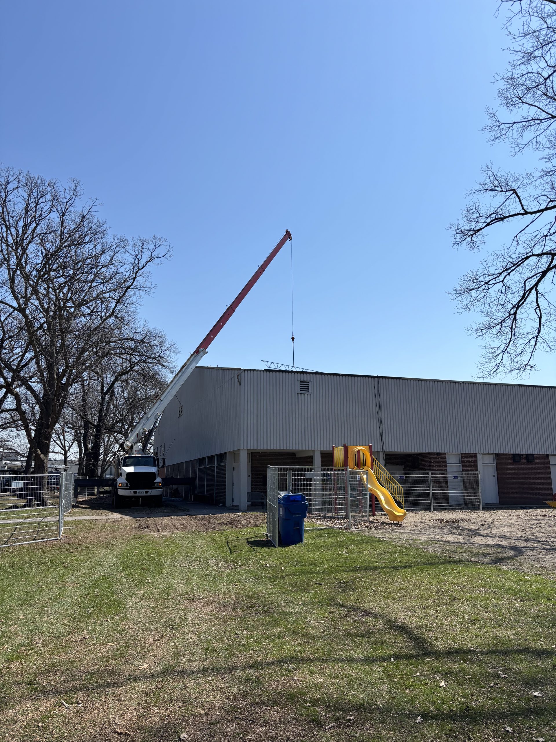 George Bell Arena viewed from the outside on a clear day. A truck with a crane is parked by the building, which is partially surrounded by safety fencing. The nearby area includes grass fields, trees and a small play structure.