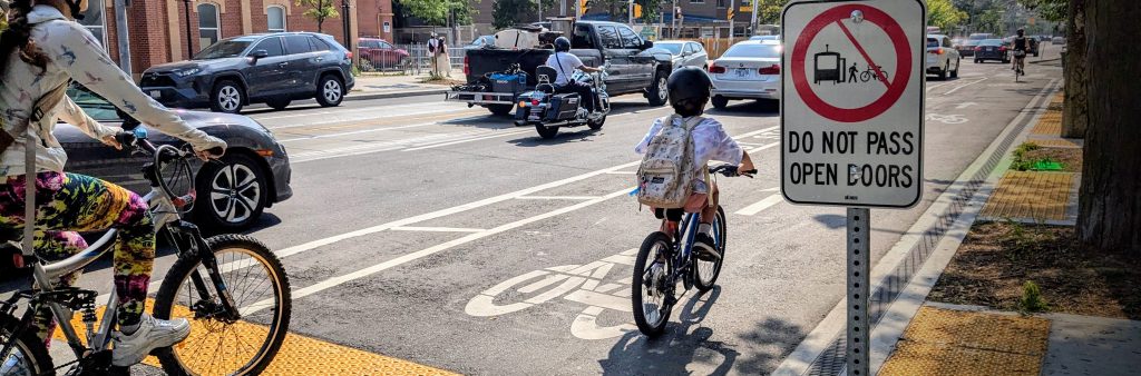 An adult and child cycle in a cycle track that is raised to sidewalk level and separated from the roadway.