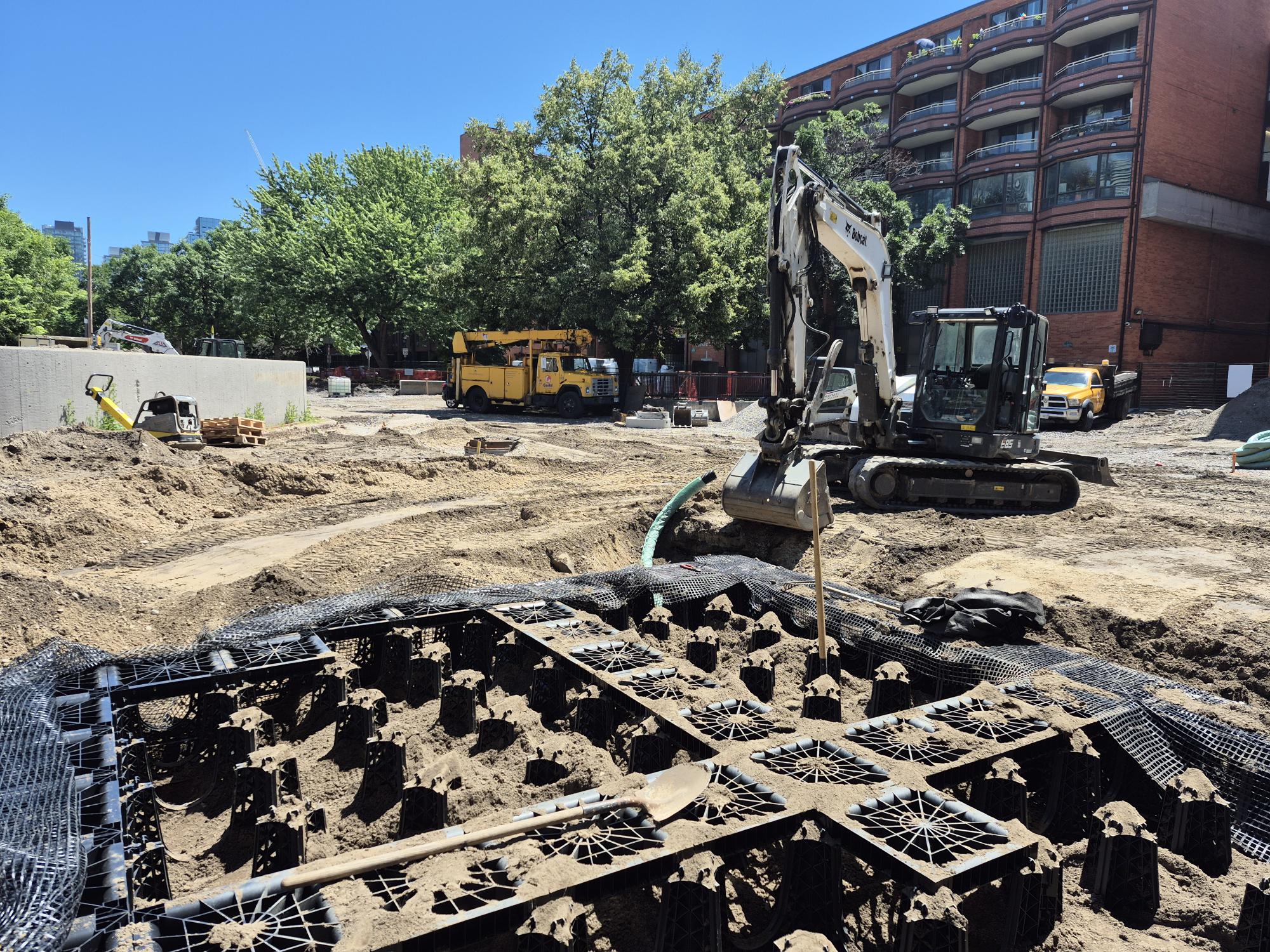 Construction site with an excavator, truck and other equipment visible. An underground installation is underway, surrounded by soil and construction materials. Mature trees and residential buildings are visible in the background.