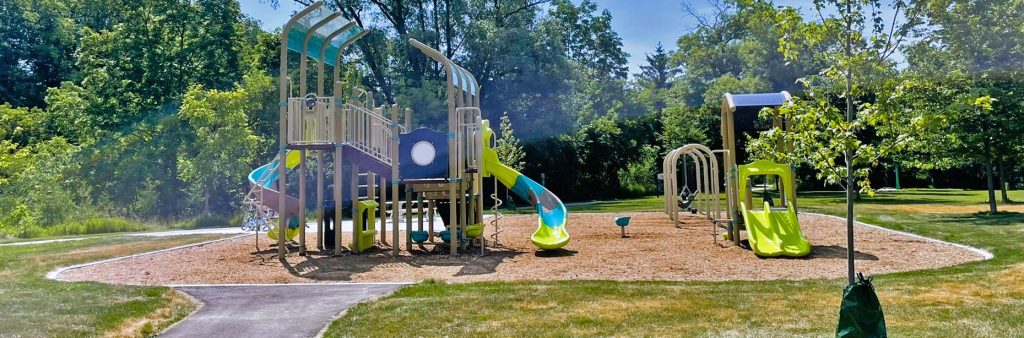 Image of the new Checkendon Park playground looking from the entry pathway towards the playground area. From left to right, the image shows the main senior play structure with a spiral slide and a curved slide, a standalone spinner, a swing set and a junior play structure with double slides. The new playground is on a woodchip surface surrounded by a grassy field with trees in the background