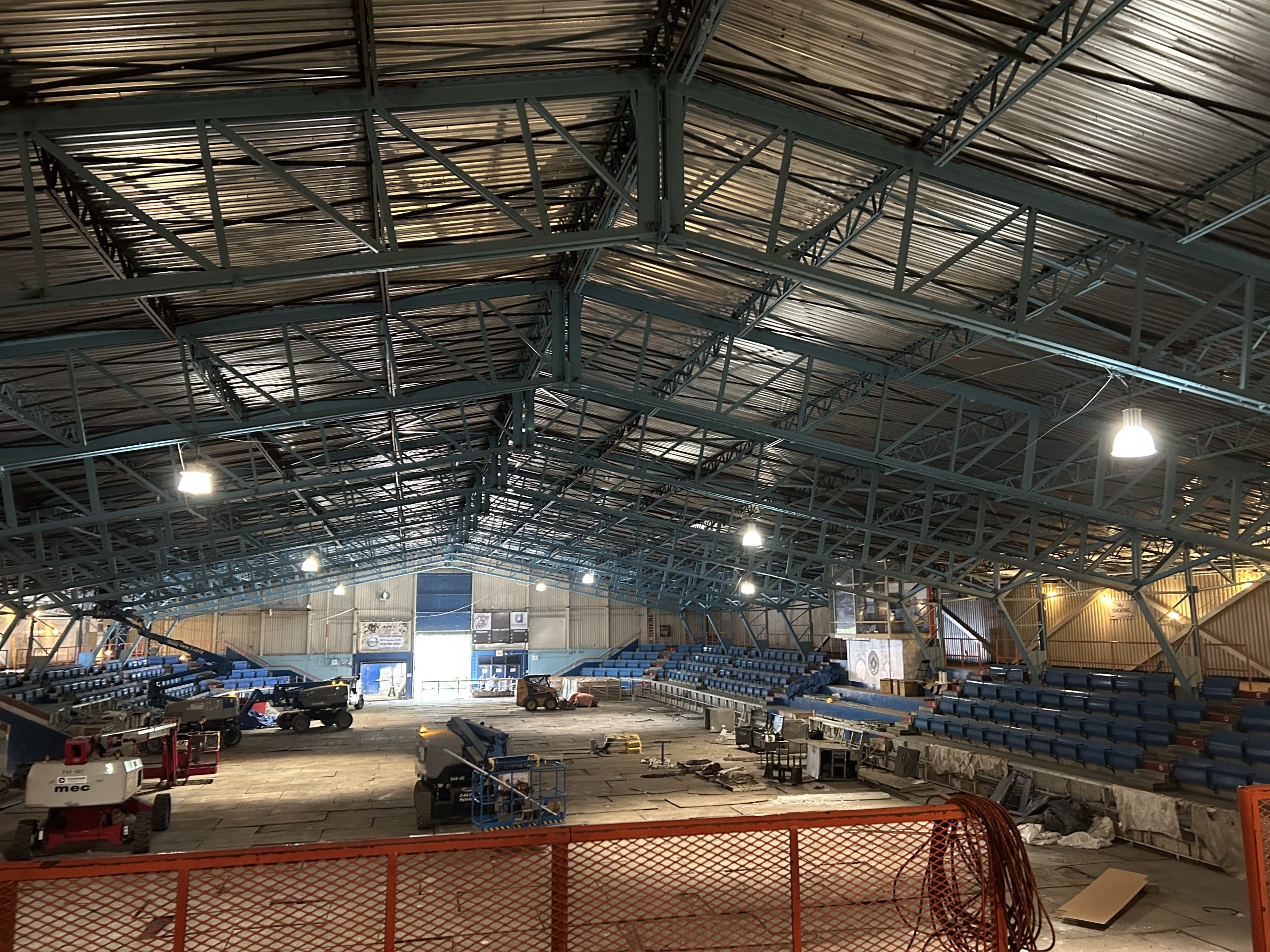 The rink in George Bell Arena under construction. Construction equipment and materials are scattered on the unfinished rink base. Rows of blue seats line the sides. The large space includes overhead lighting and exposed roof trusses.