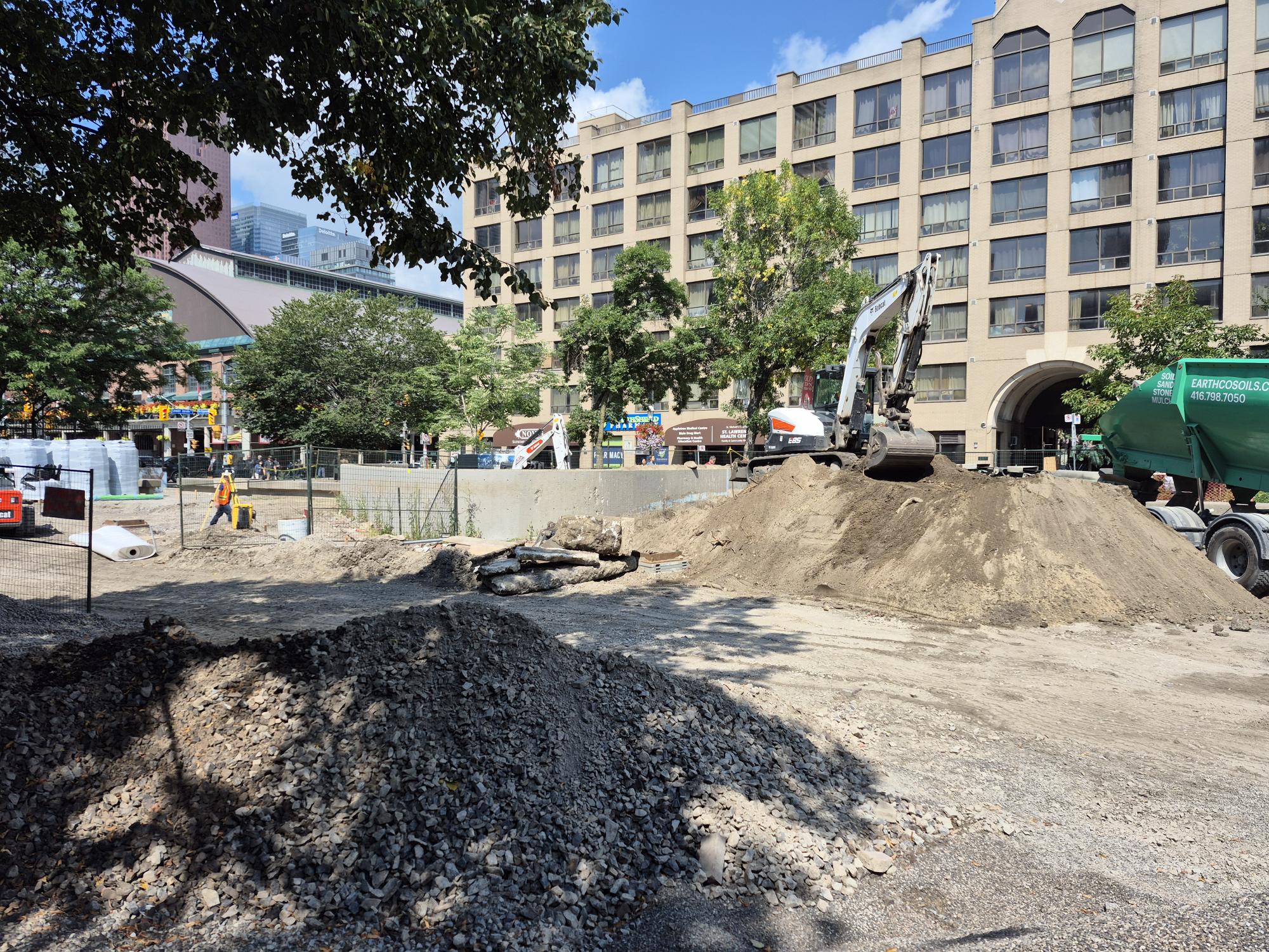 Construction site with piles of gravel and soil, excavators and temporary fencing. A worker is visible onsite, while apartment buildings, mature trees and stores line the background.