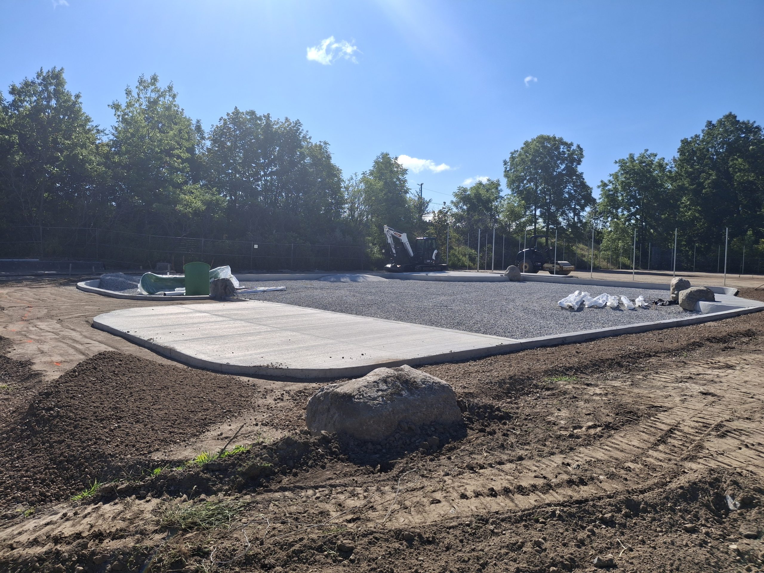 Outdoor construction site in a park, featuring a curved area filled with gravel and connected to a concrete pad. Large rocks are placed around the area, which is surrounded by dirt. The entire site is bordered by chain-link fencing, and construction machines are visible in the background.