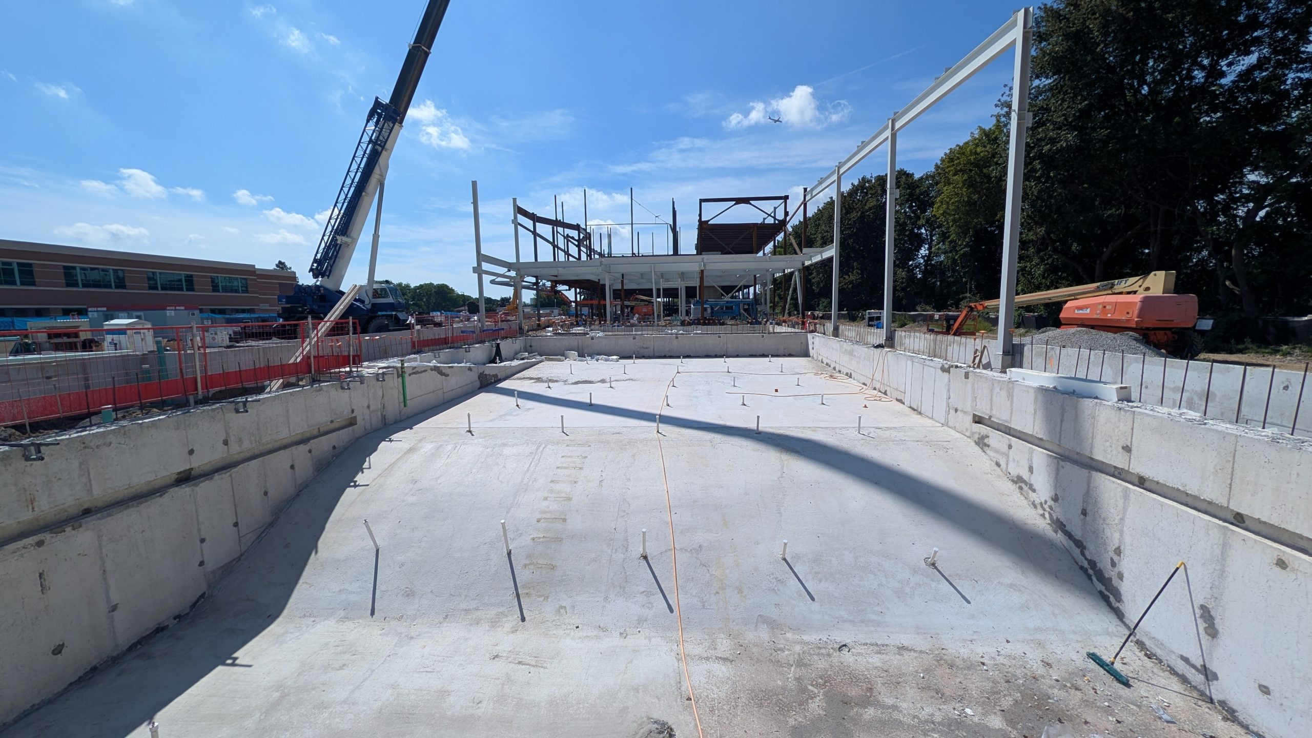 View of the construction site for the new community recreation centre, showing a large concrete foundation area with utility connections visible across the slab. Steel columns are being installed around the perimeter, while a crane, lifts and an unfinished building structure are visible in the background.