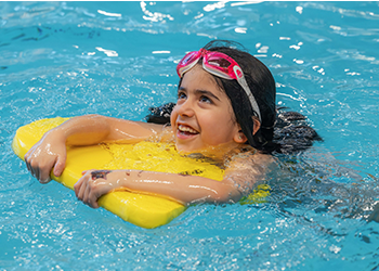 A girl swimming in a pool with a yellow kickboard and pink goggles.