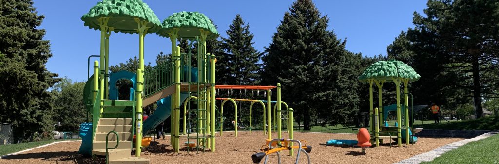 view of playground from the new concrete walkway to the south-west, looking north-east. Senior play structure and tetter totter in the foreground, junior structure to the back right, and swings in the middle background.
