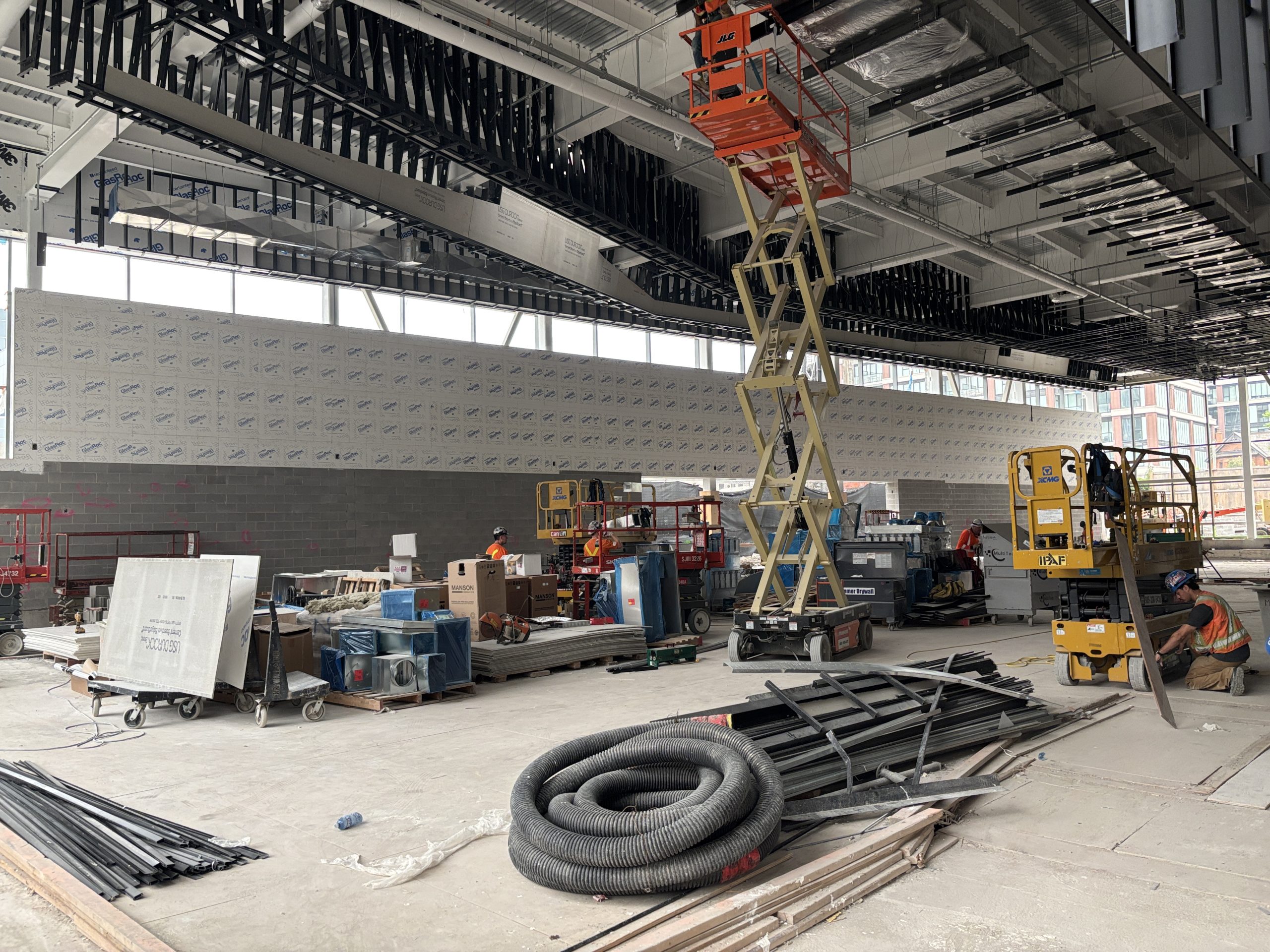 Interior view of Davisville Community and Aquatic Centre, showing a large space under construction with high ceilings, exposed framing and ducts. A partially tiled wall lines one side of the room, while large windows line another side. Construction materials and equipment, such as coiled conduits and scissor lifts, are scattered around the site, with workers in safety vests visible.