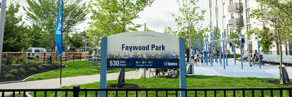 Park sign at the entrance of Faywood Park. A paved pathway near the entrance leads to a play area with climbing structures and benches, surrounded by grass and trees. Community members are seen enjoying the park.