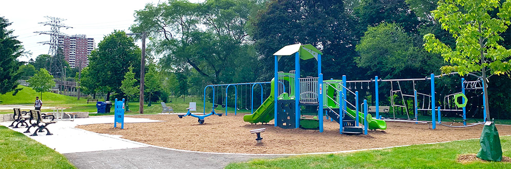 New Prairie Drive Park playground looking south-west from the redone walkway. Muskoka chairs to the left, swing to the left, climbing structure in the centre and junior play structure with individual play elements to the right. Washrooms building and mature trees in the background.