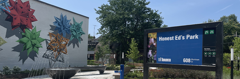 The new park on a clear day, featuring a decorative water feature, a drinking water fountain, trees, plantings and paved paths. In the foreground, the park sign reads “Honest Ed’s Park.” A bright mural is painted on the wall of the building next to the park.