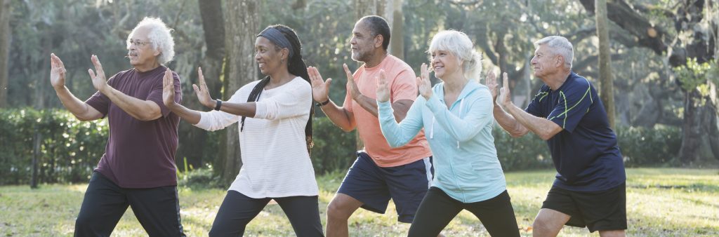 Five older adults practices tai chi together in a park, standing in a staggered formation with their arms extended in a slow, synchronized movement.