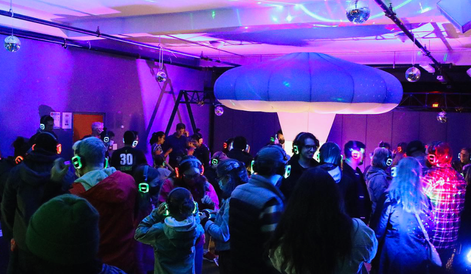 Crowd wearing neon light up headphones in a room with mushroom installation, disco balls and light projected onto the ceiling