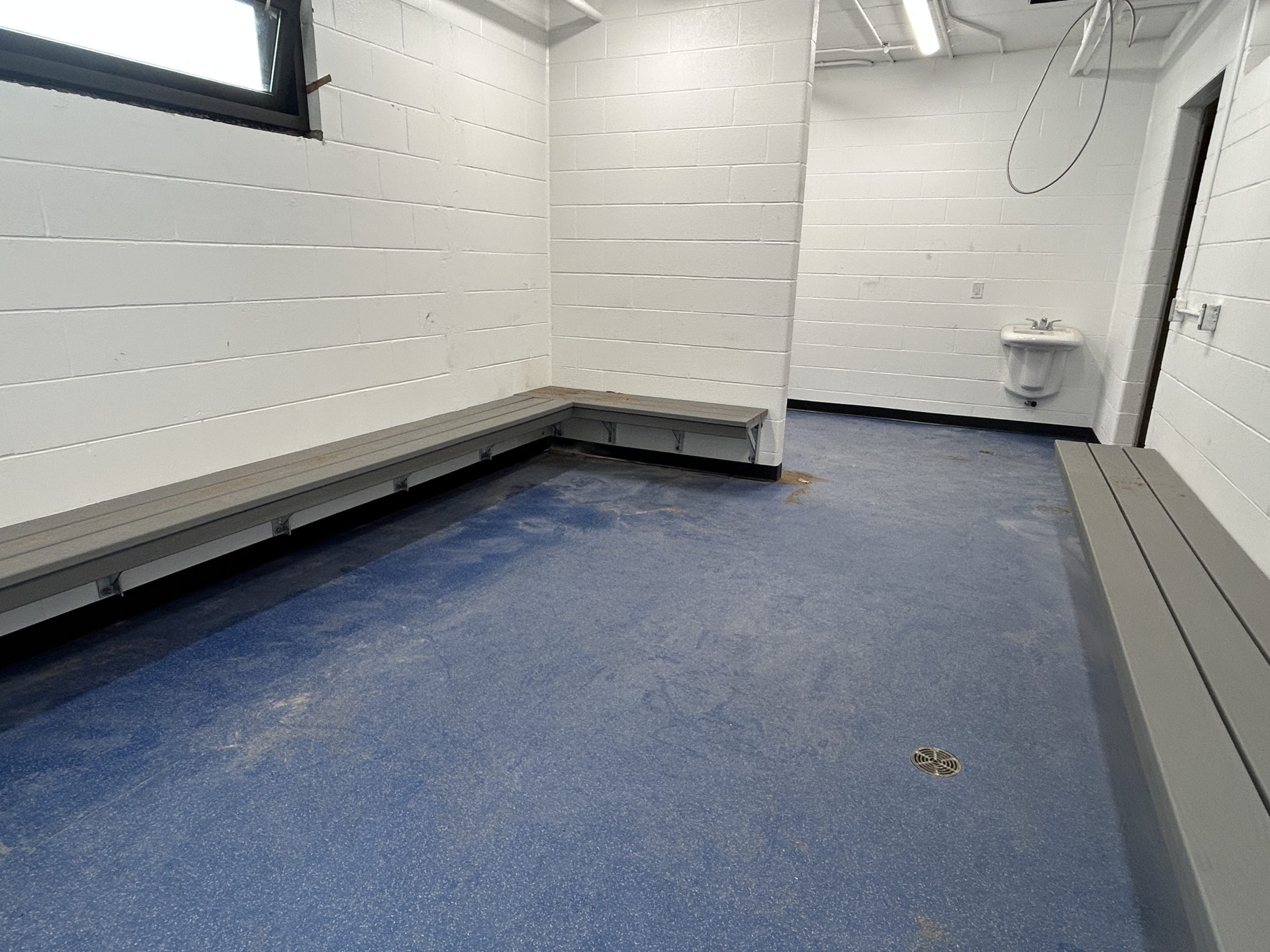 A dressing room in George Bell Arena with grey benches installed along white block walls and blue speckled flooring. Overhead lighting and natural light from a window illuminate the space. A small sink is mounted on the far wall.