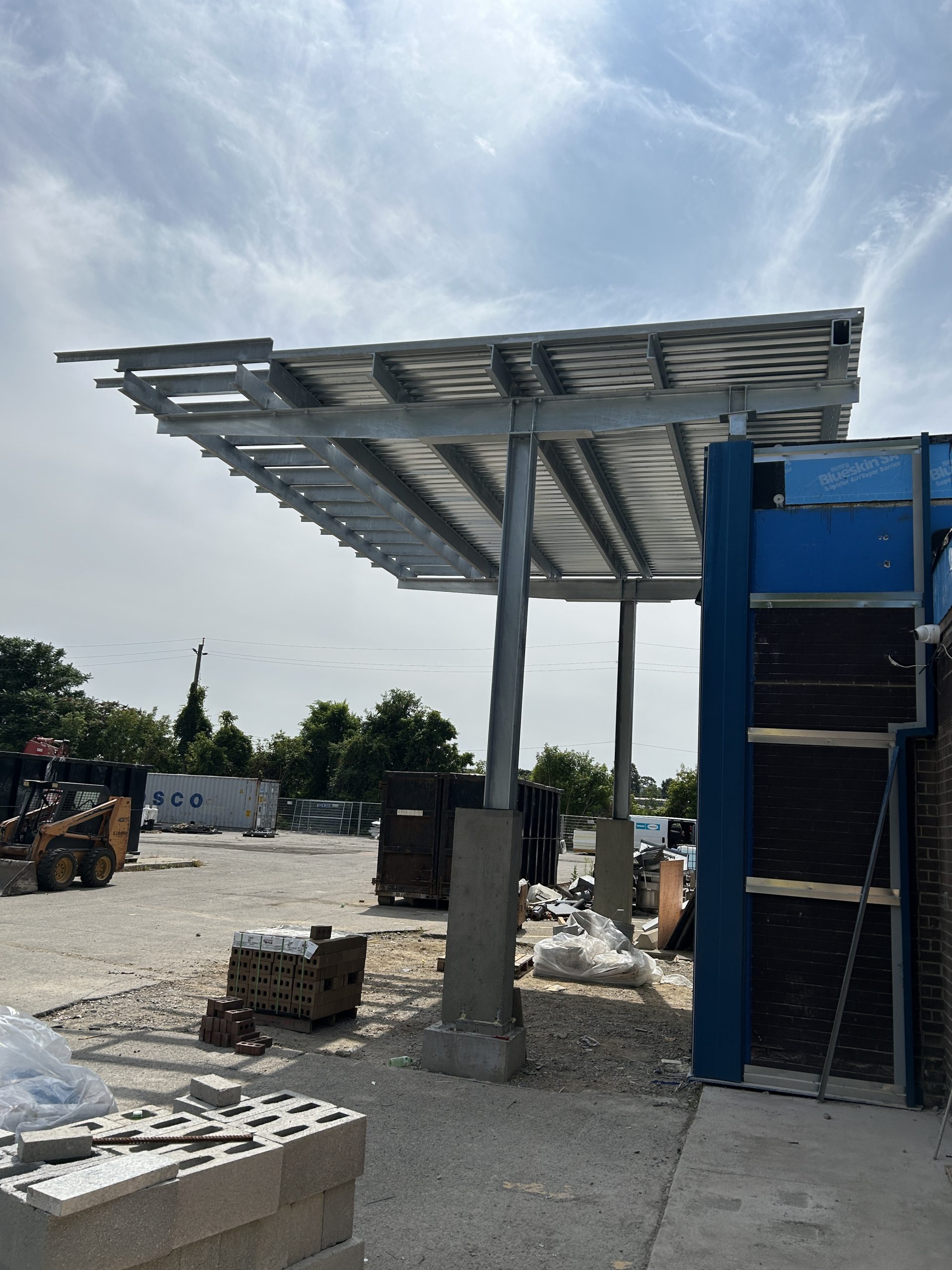 A steel entrance canopy attached to the exterior of a building. Construction materials and equipment are visible in the surrounding lot.