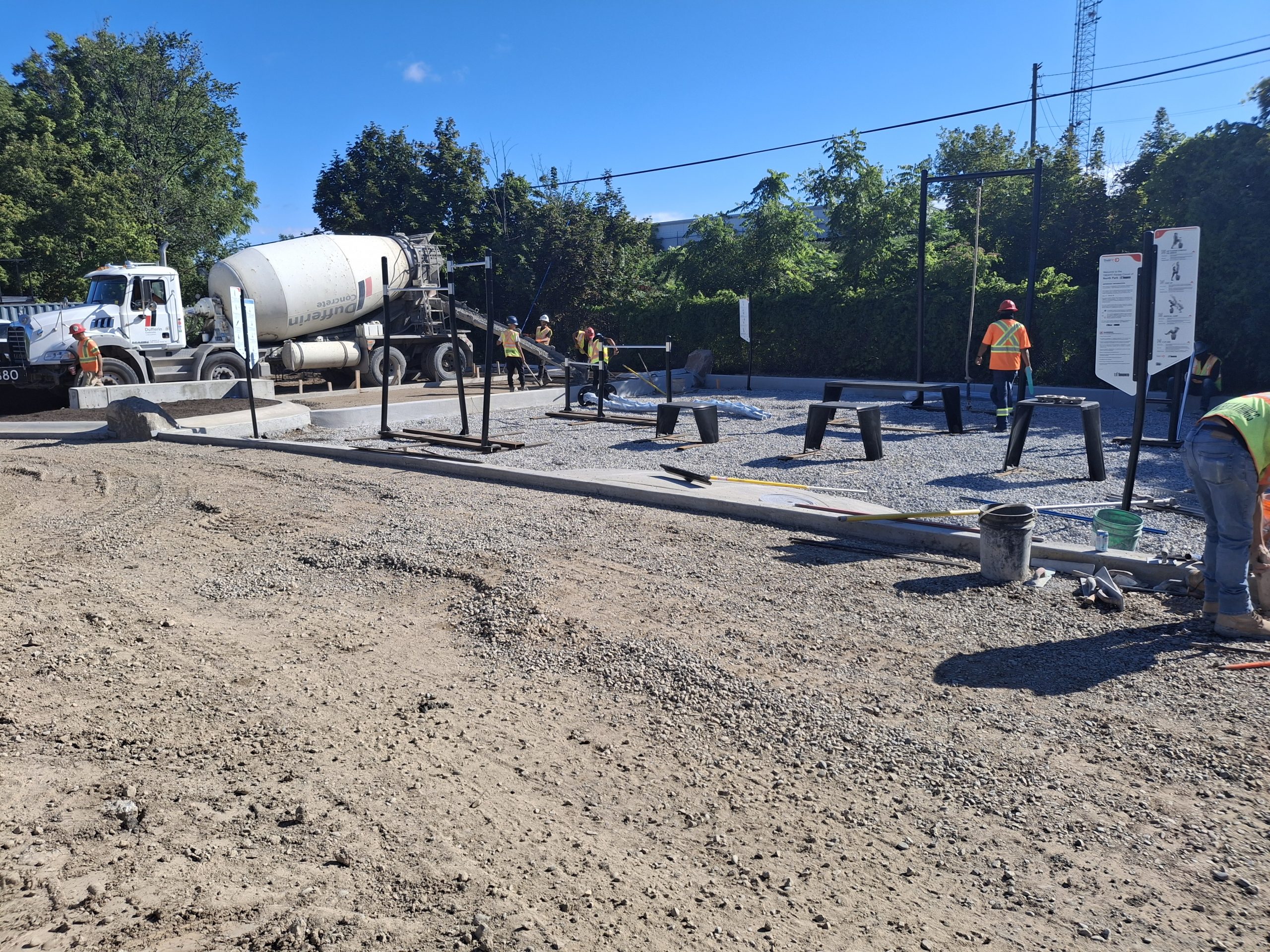 Ongoing construction for an outdoor fitness area. Fitness equipment, such as pull-up bars and step benches, and instructional signs are installed in a bed of gravel. Construction workers, materials and equipment are visible around the site, including a concrete mixer truck.