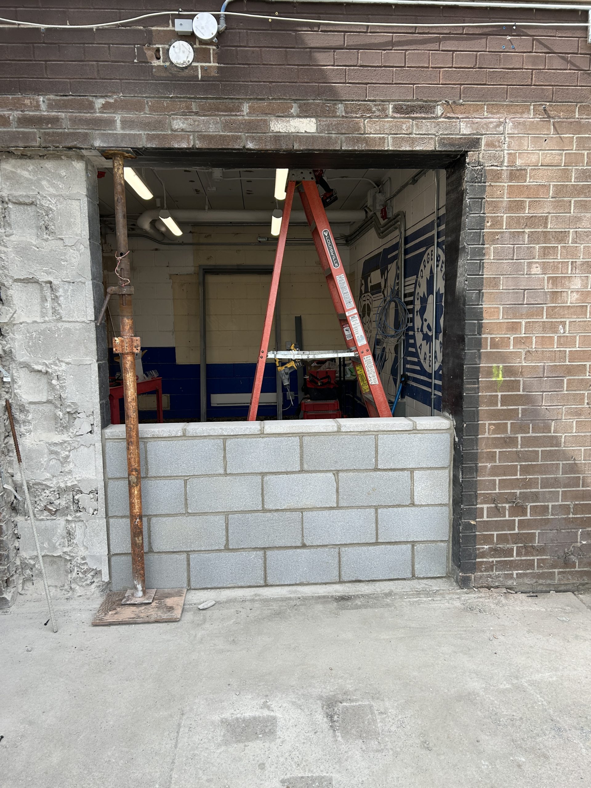 A brick wall with a partially enclosed opening through which an indoor arena space with painted walls is visible. Concrete blocks have been installed along the bottom half of the opening.