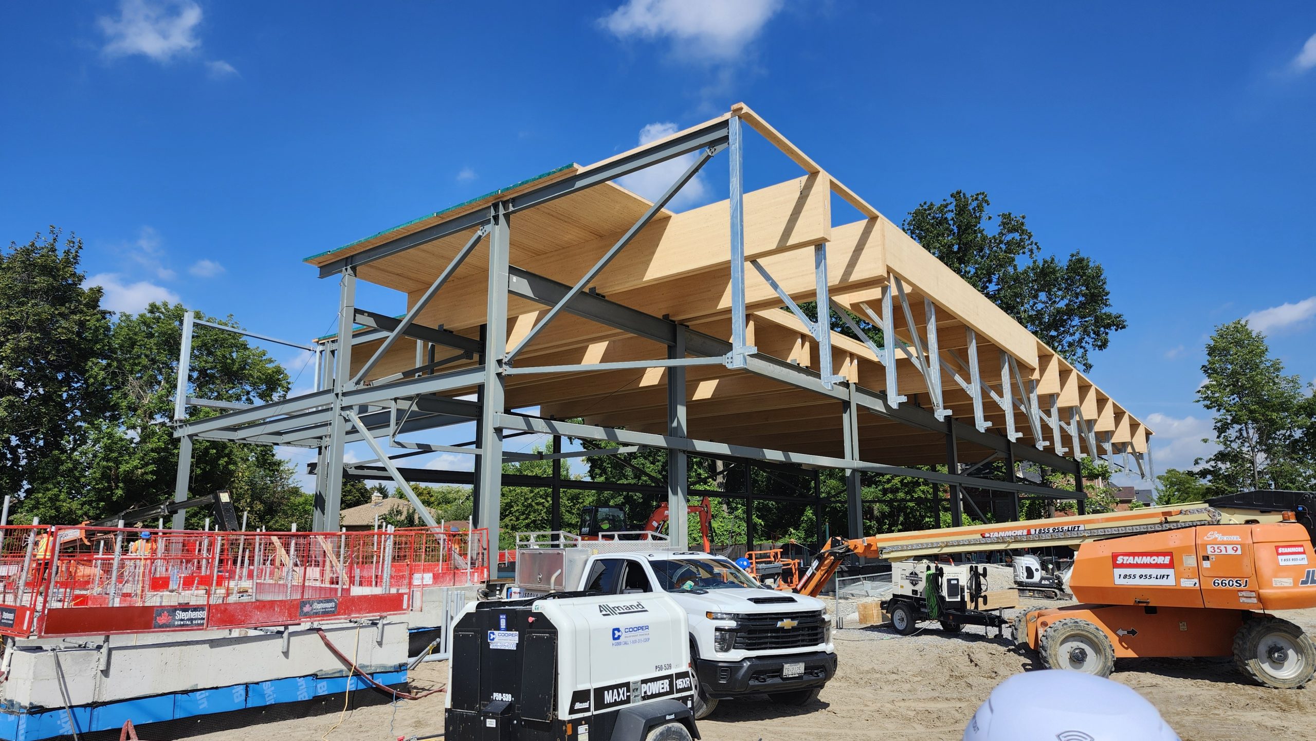 View of the construction site for the new community recreation centre, showing the installation of timber panels on top of a steel building frame. A construction lift, excavator and other equipment are visible around the site, along with workers in safety vests.