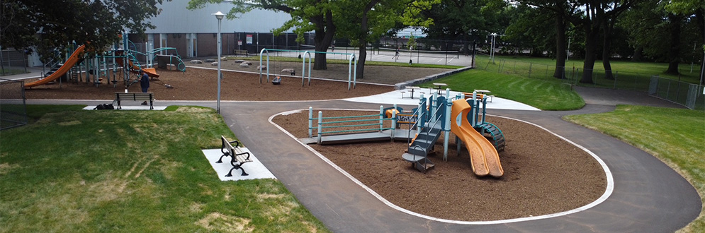 Aerial view of a park playground featuring two play structures on separate wood chip surfaces connected by winding pathways. The surrounding area includes grass lawns, benches, picnic tables and mature trees. Fenced sports courts are visible in the distance.