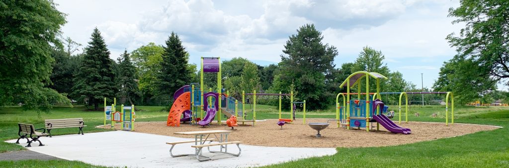 New playground area looking south from the open lawn. Seating area with two benches and picnic table on concrete surfacing in the foreground, with the playground with wood fibre surfacing in the middle ground, mature trees and open lawn in the background.