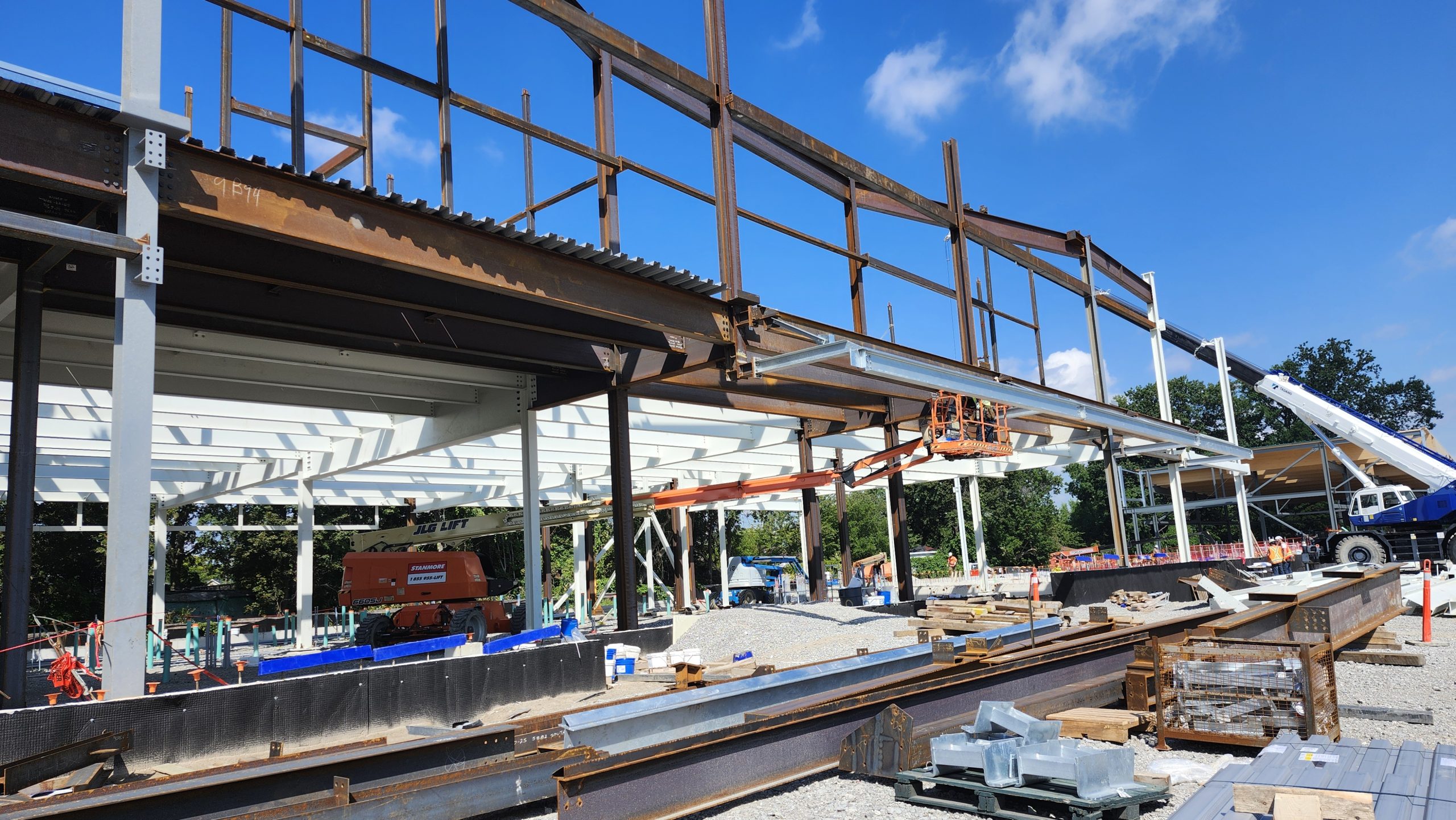 View of the construction site for the new community recreation centre, showing the erection of structural steel framework. The framework consists of rust-coloured and white vertical and horizontal beams. Workers in a lift are installing a beam. Steel materials and construction supplies are piled in the foreground.